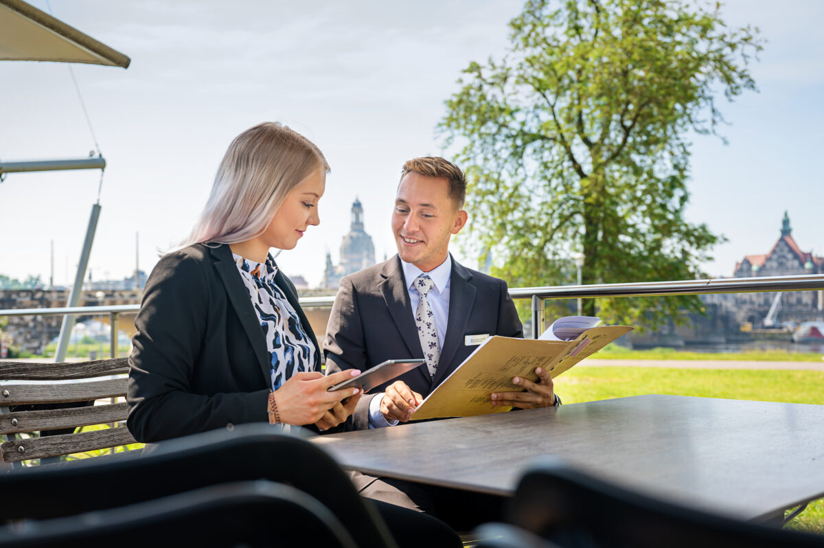 An der Terrasse sitzen zwei Menschen und schauen ein Dokument.