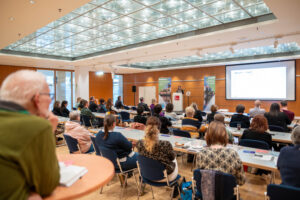 Viele Menschen sitzen in einem Raum zu Beginn der landesweite TI-Informationsveranstaltung.